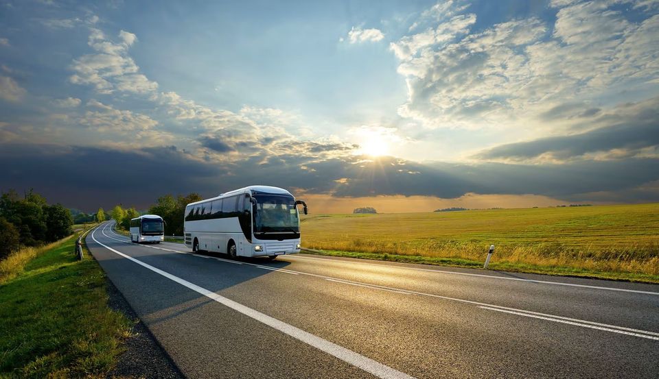 Two white tour buses drive along a highway through a scenic countryside under a partly cloudy sky. Two white tour buses drive along a highway through a scenic countryside under a partly cloudy sky.